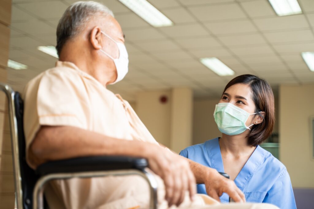 Asian nurse taking care of mature male patient sitting on wheelchair in hospital. Young woman and old man wearing surgical face mask for protection of The COVID pandemic. Girl talking to elderly man.